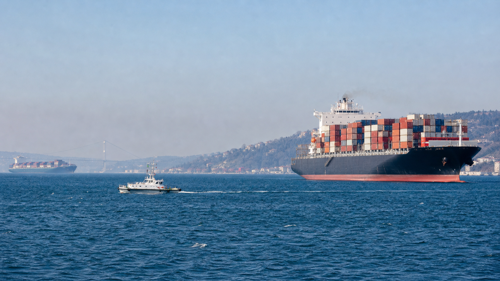 Commercial container ships and small patrol boats navigating a wide shipping channel at sea under clear daylight, with distant coastline visible.