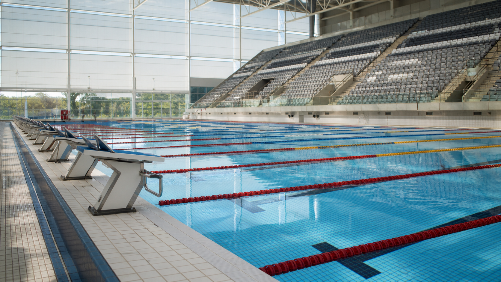 Competition swimming pool lanes and starting blocks with an empty laneside area and spectators in the stands visible in soft light.
