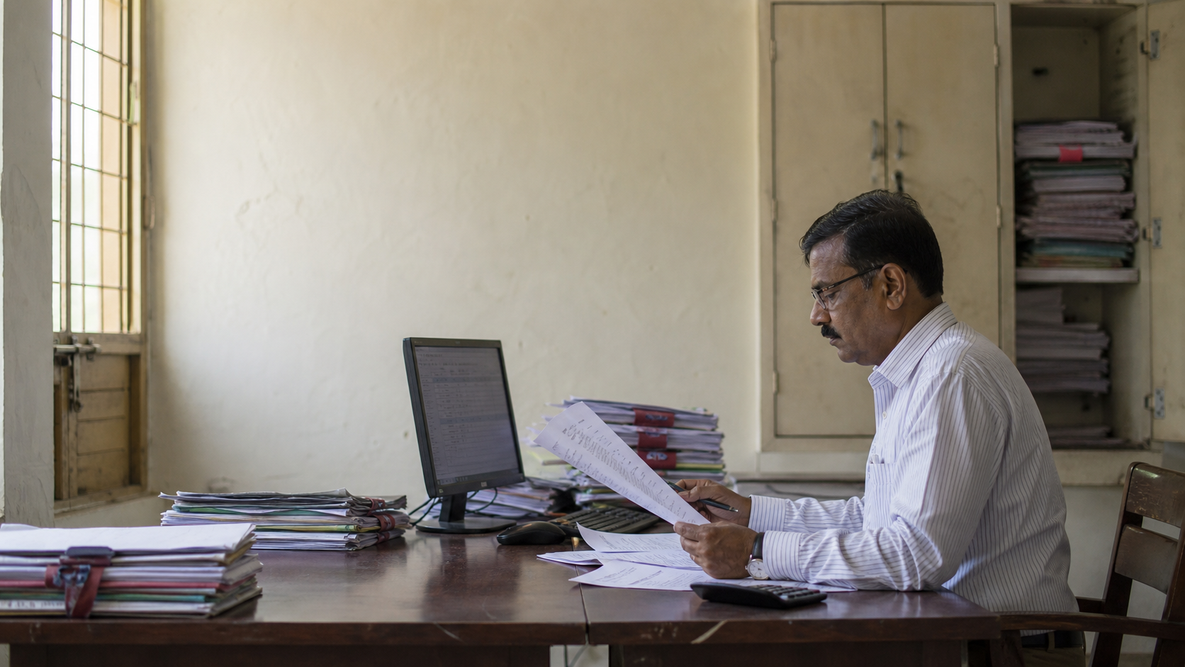 A state treasury clerk in India reviews daily cash-flow sheets beside a desktop terminal as sunlight falls across stacked vouchers and sealed envelopes.