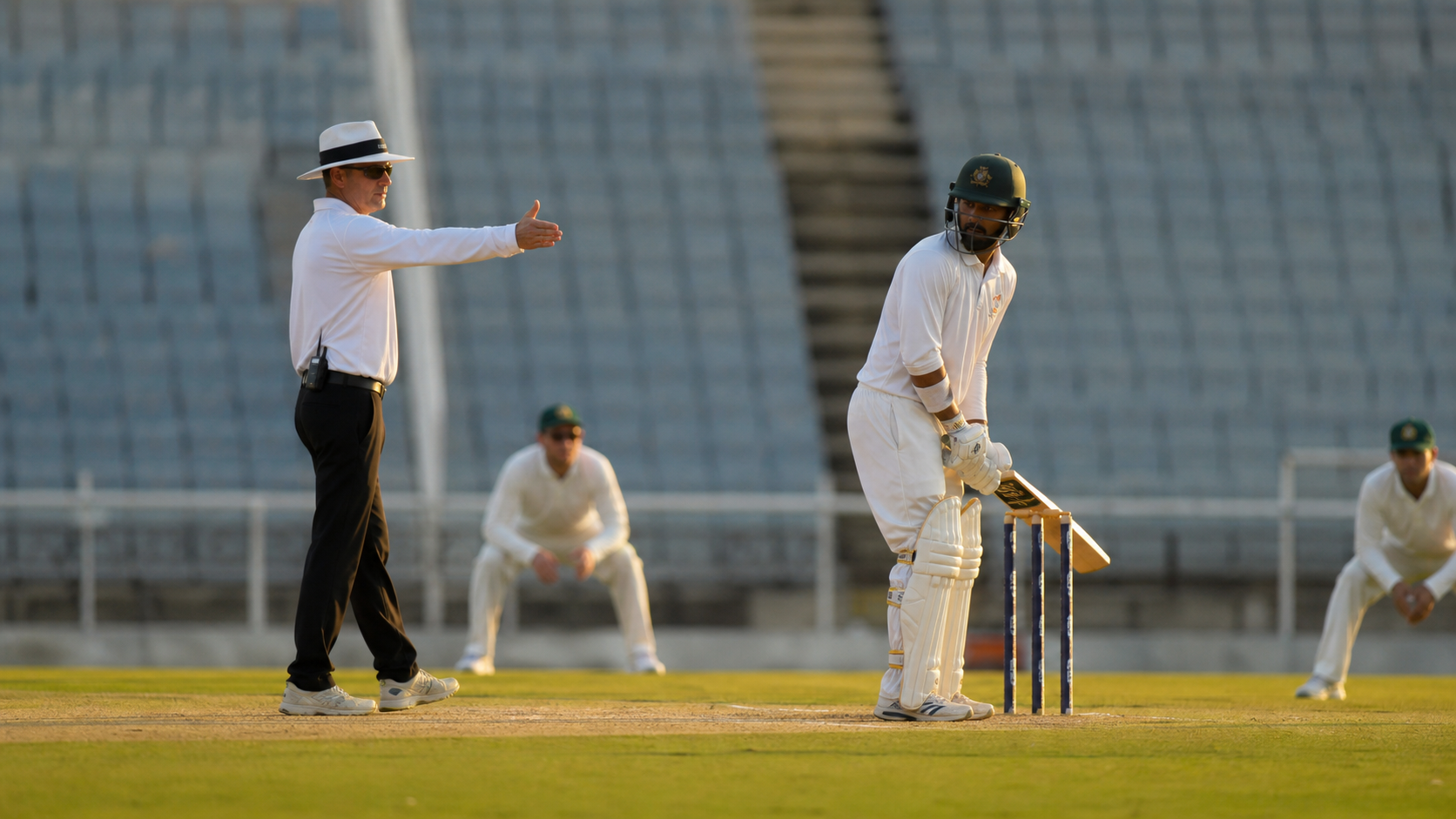 Umpire signaling to the scorer while a batter adjusts stance at the crease under late-afternoon light, with fielders waiting and a blurred scoreboard in the distance