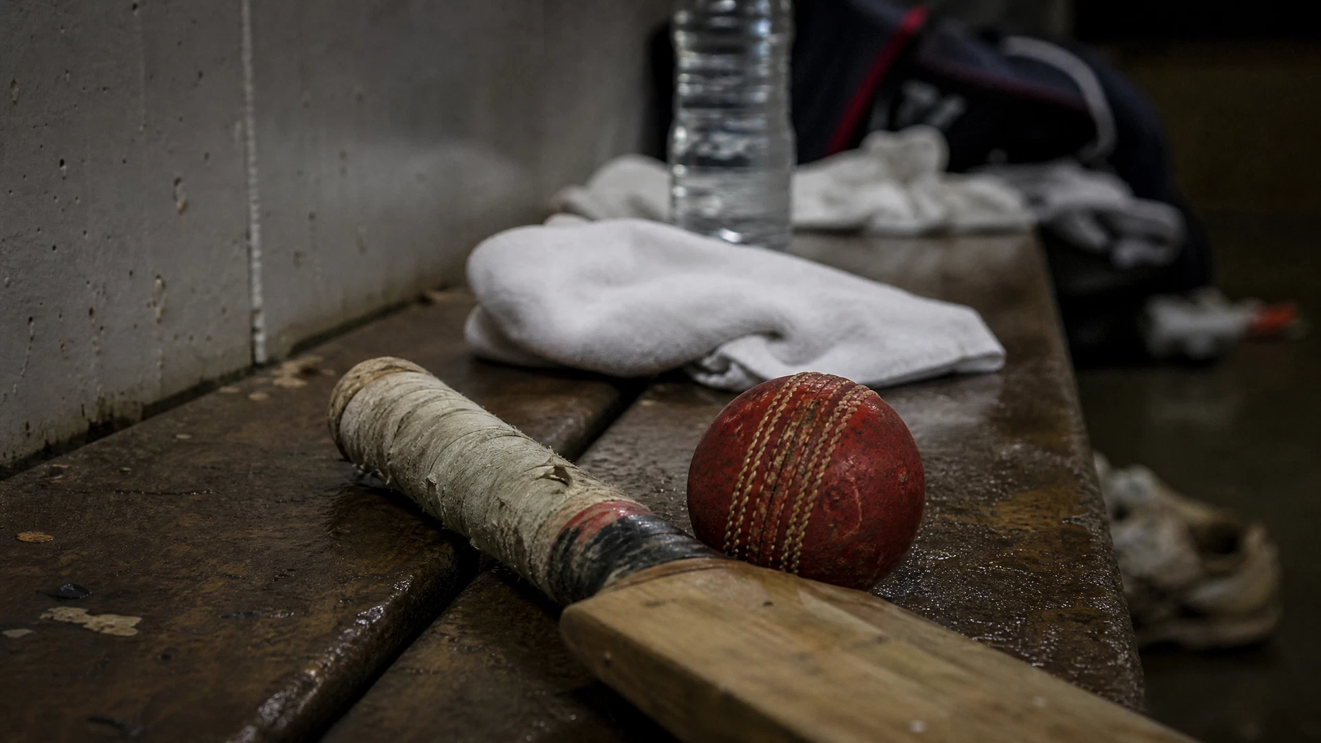 A worn cricket ball and taped bat rest on a scuffed changing-room bench after training.