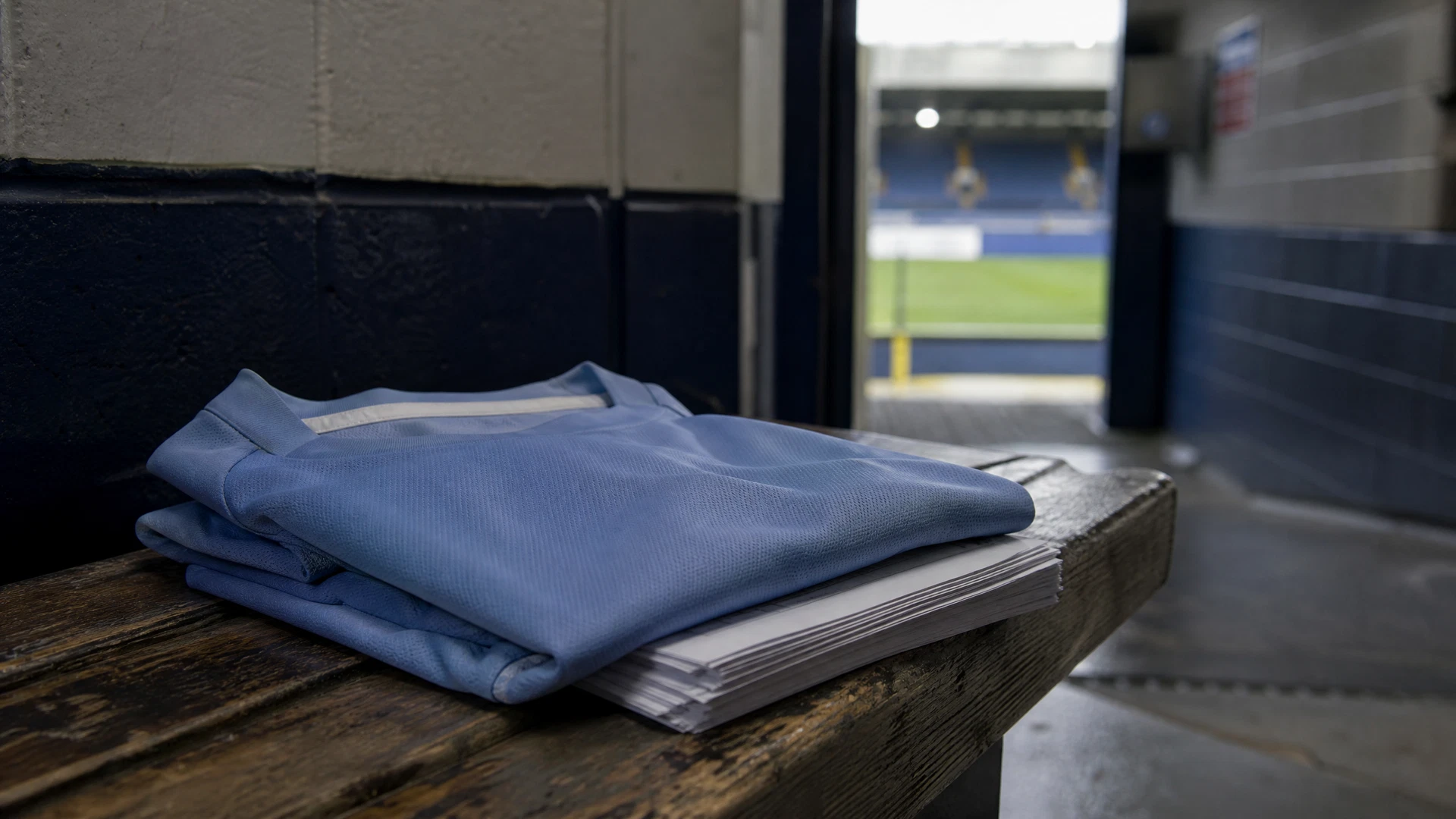 A folded light-blue jersey and unsigned paperwork rest on a worn changing-room bench.