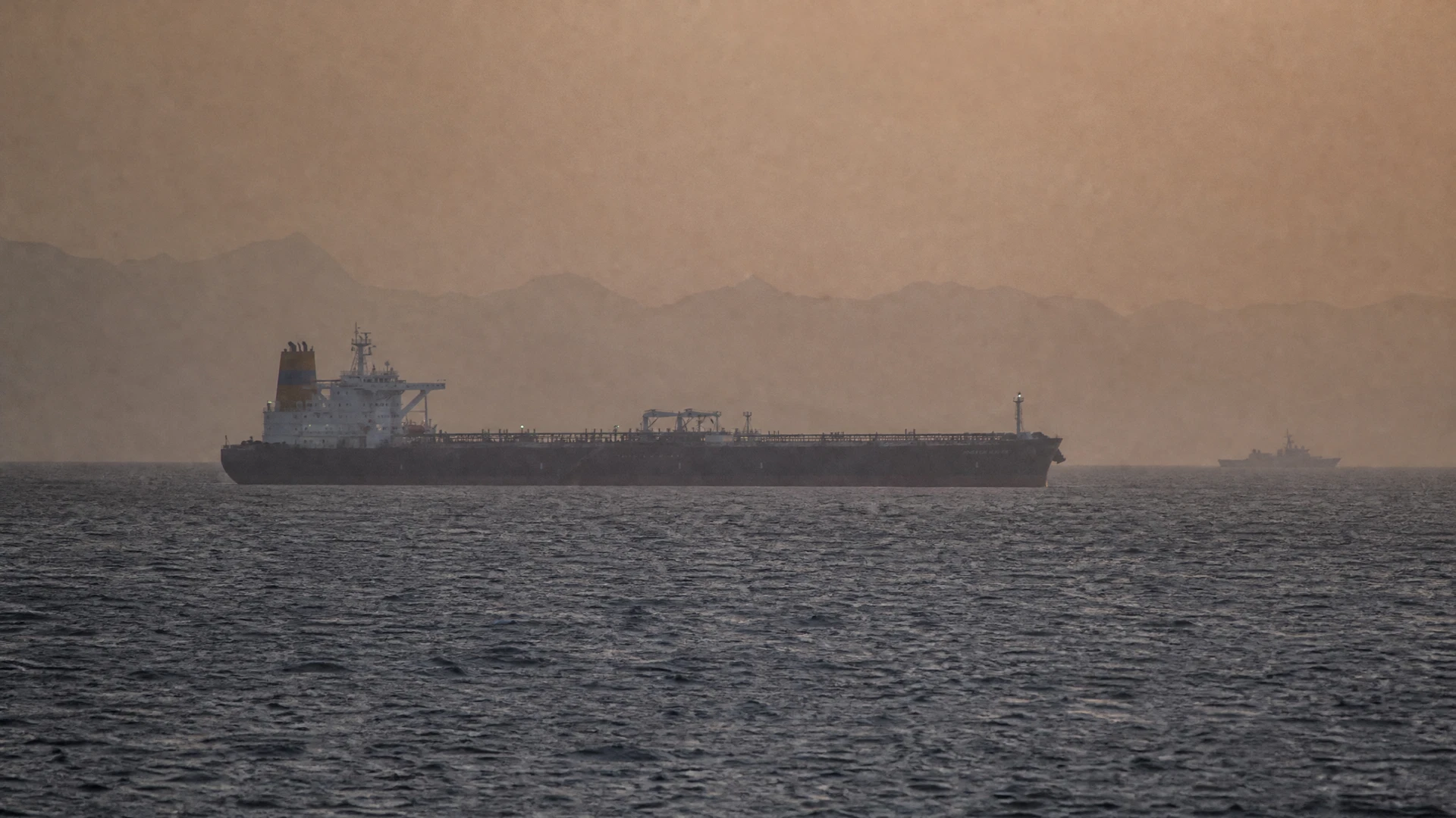 A commercial oil tanker crosses the hazy Strait of Hormuz with a distant patrol vessel astern.