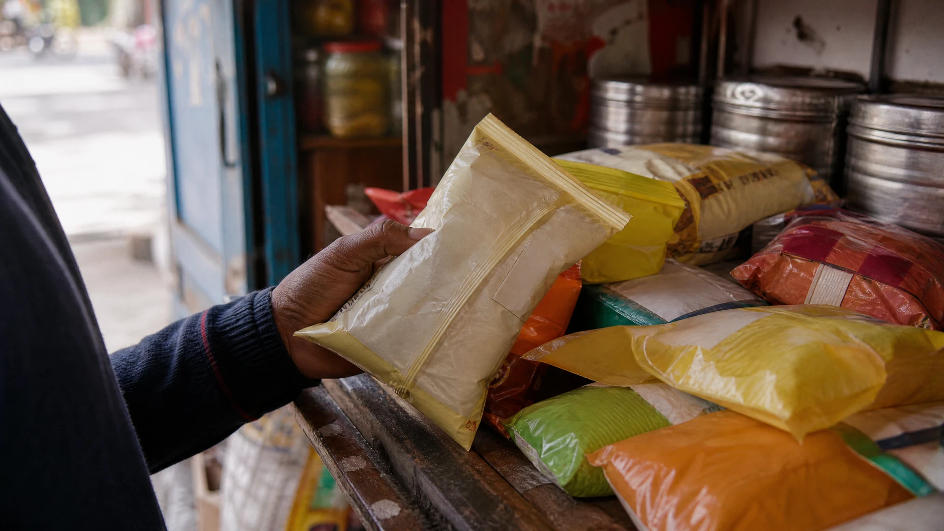 A shopper examines an unbranded snack packet on a weathered neighborhood shop counter.