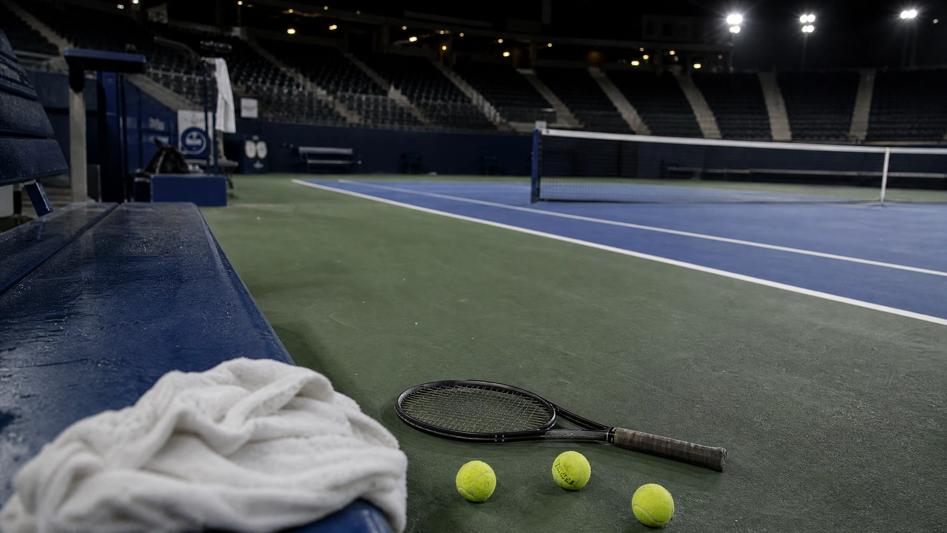 A lone racket and used tennis balls remain on an empty blue hardcourt after a night match.