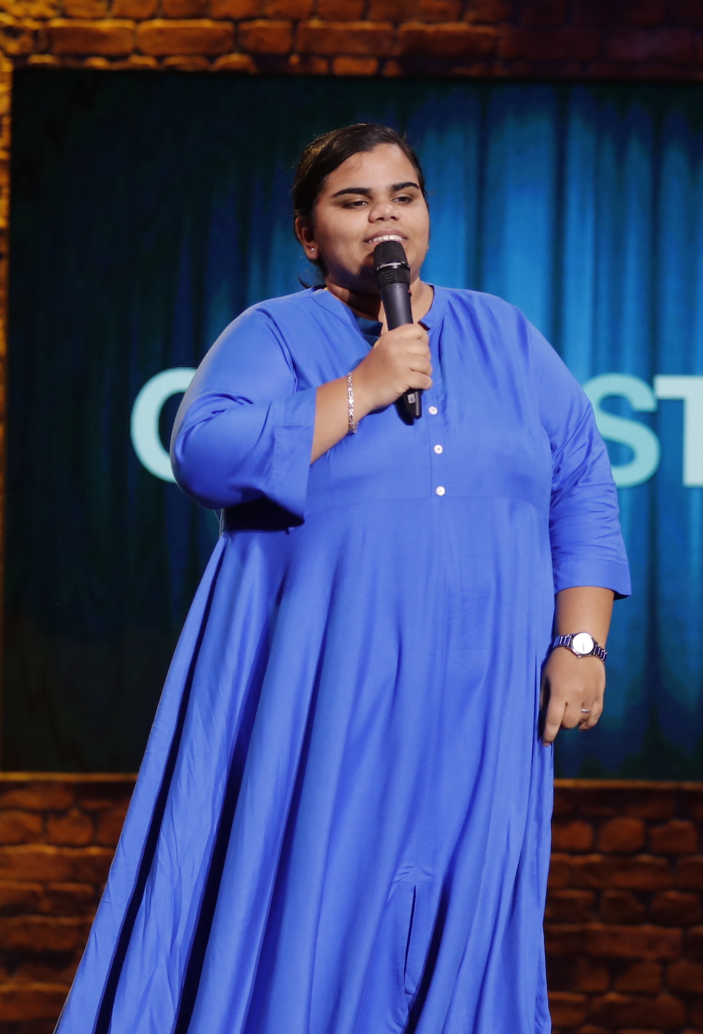 Syama Harini, a female comedian, standing alone on a stage in a blue dress and holding a microphone.