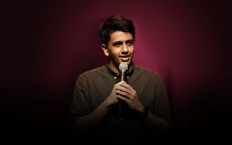 Gautham Govindan, a male stand-up comedian, holding a microphone on a dim stage against a burgundy background.