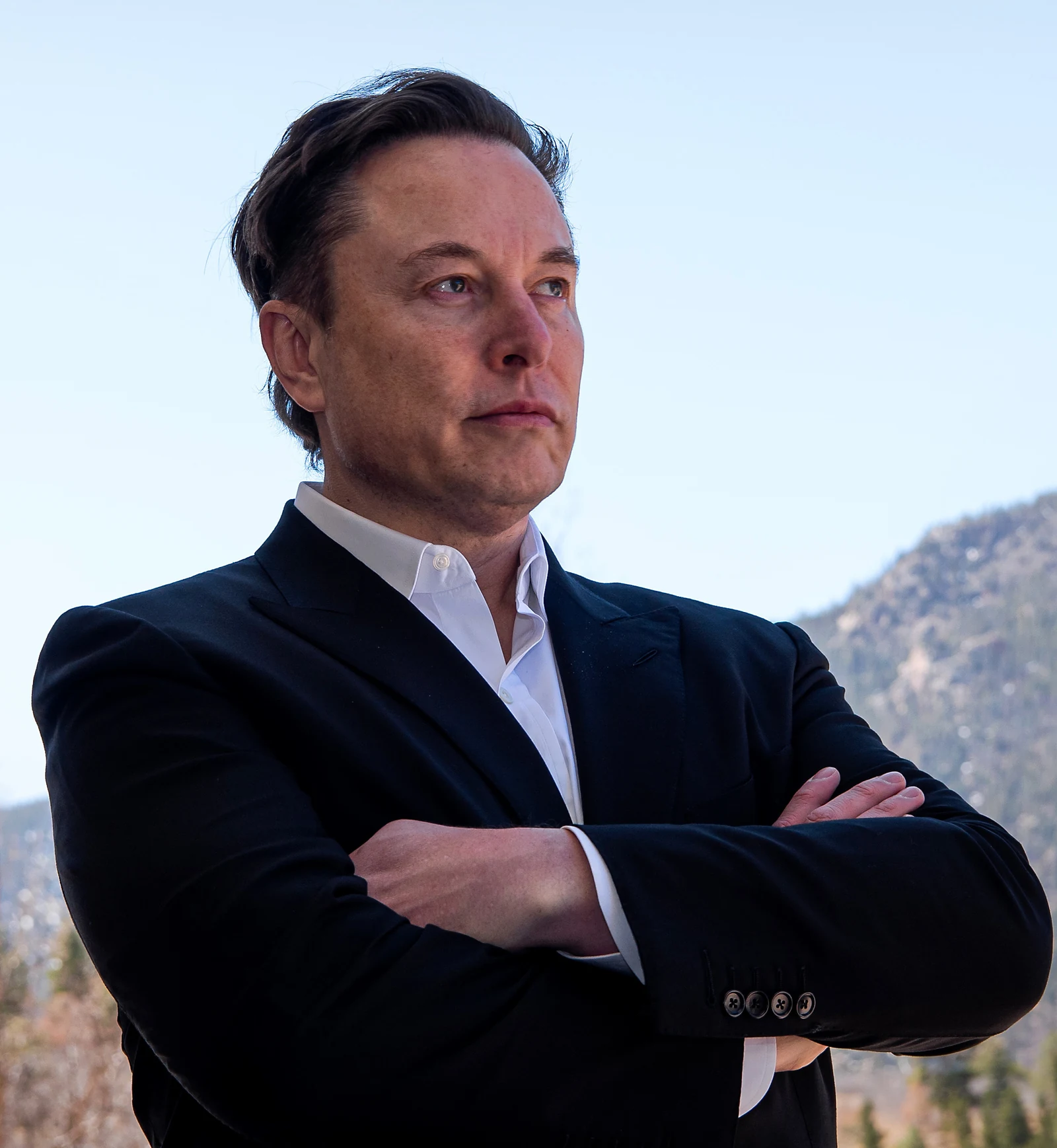 Elon Musk in a dark blazer standing outdoors with arms crossed against a mountain backdrop.