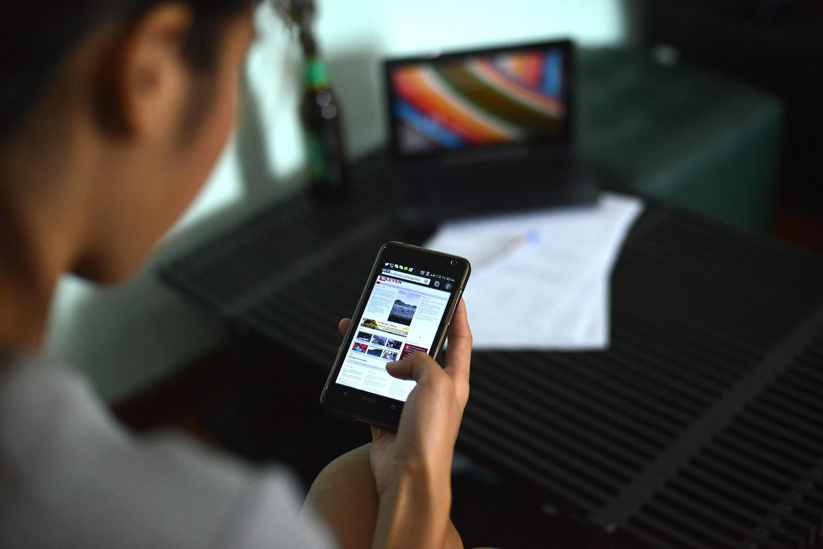 Person reading a news webpage on a smartphone while seated at a laptop indoors.
